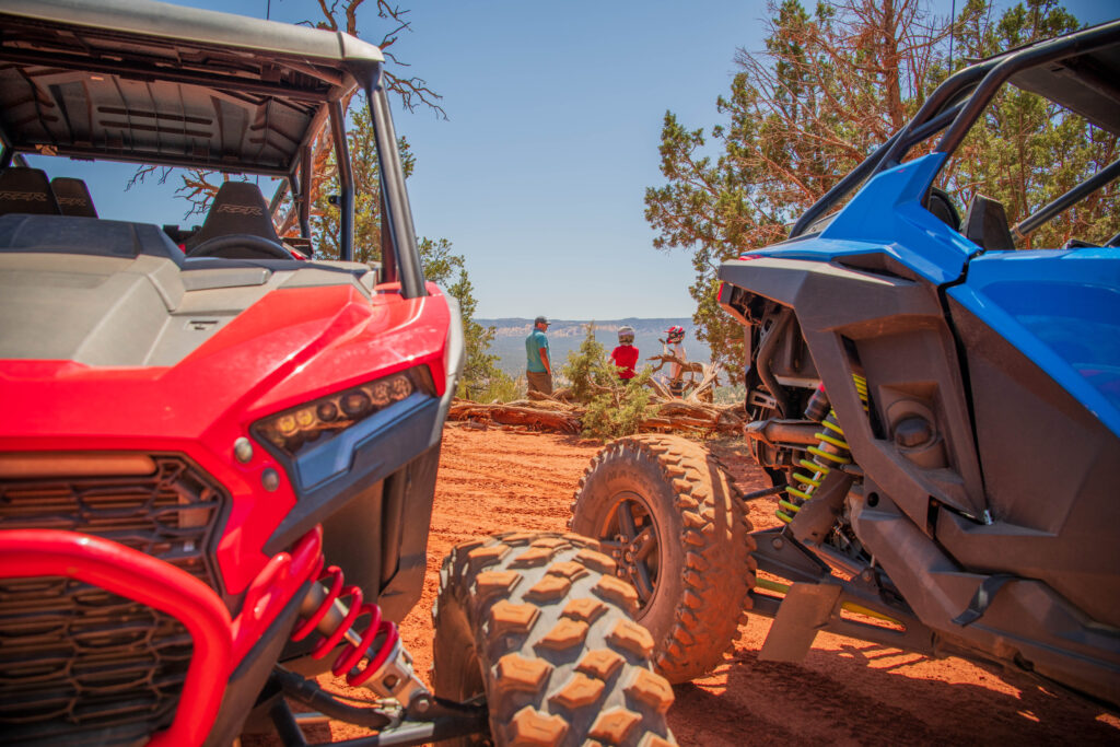 Red and blue ATVs parked on a scenic dirt trail in Kanab, Utah, with family members exploring desert trails in the background.